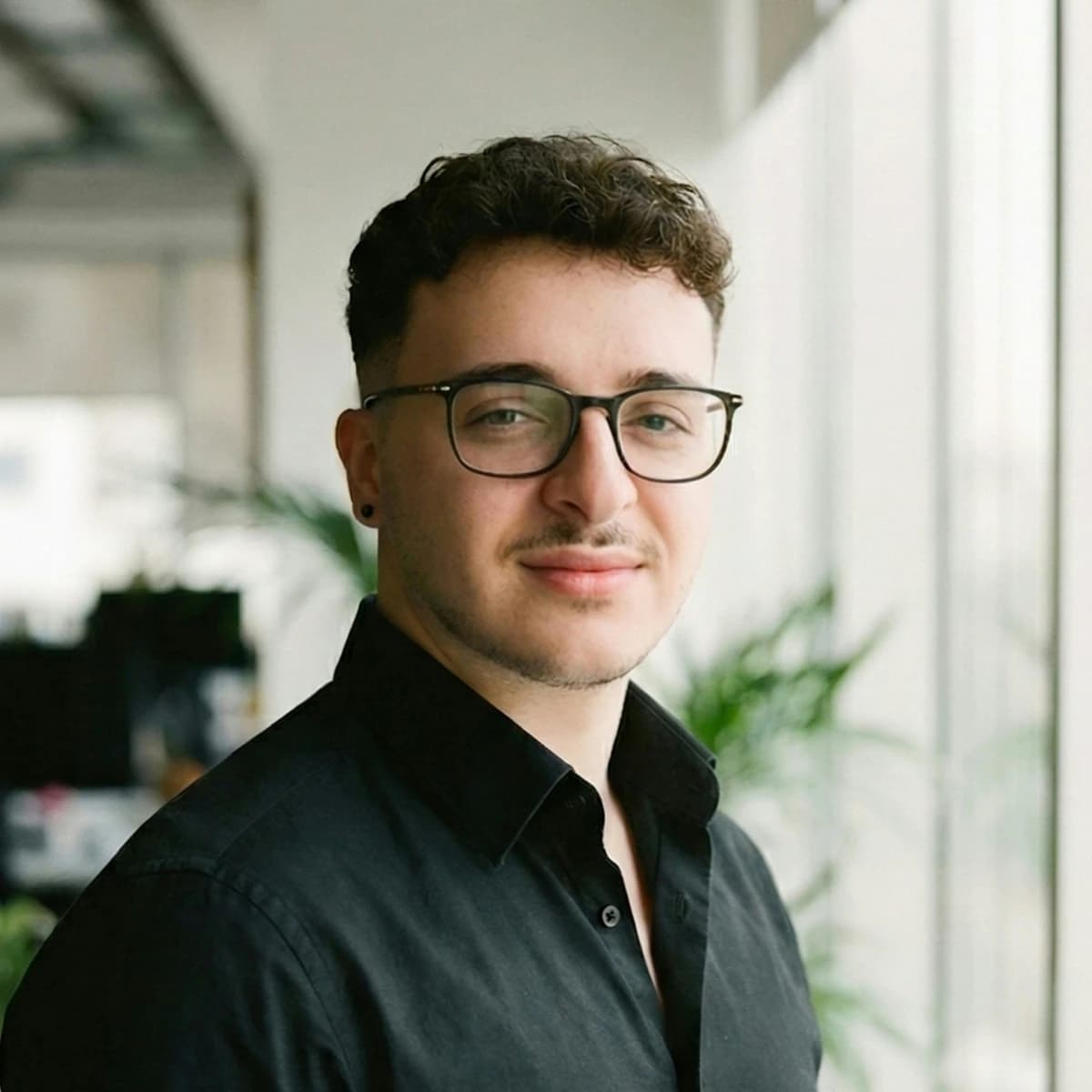 Ein Mann mit kurzen dunklen Locken und schwarzer Brille, in einem schwarzen Hemd, in einem Großraumbüro
